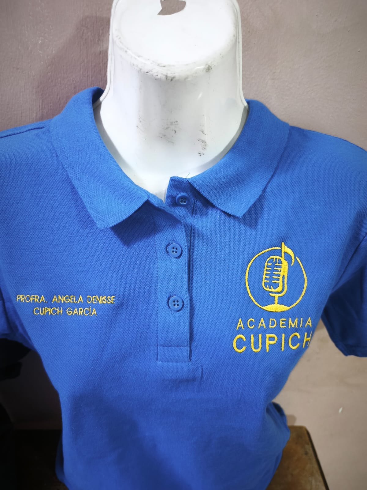 Detalle de logotipo y nombre bordados en hilo dorado sobre polo azul rey