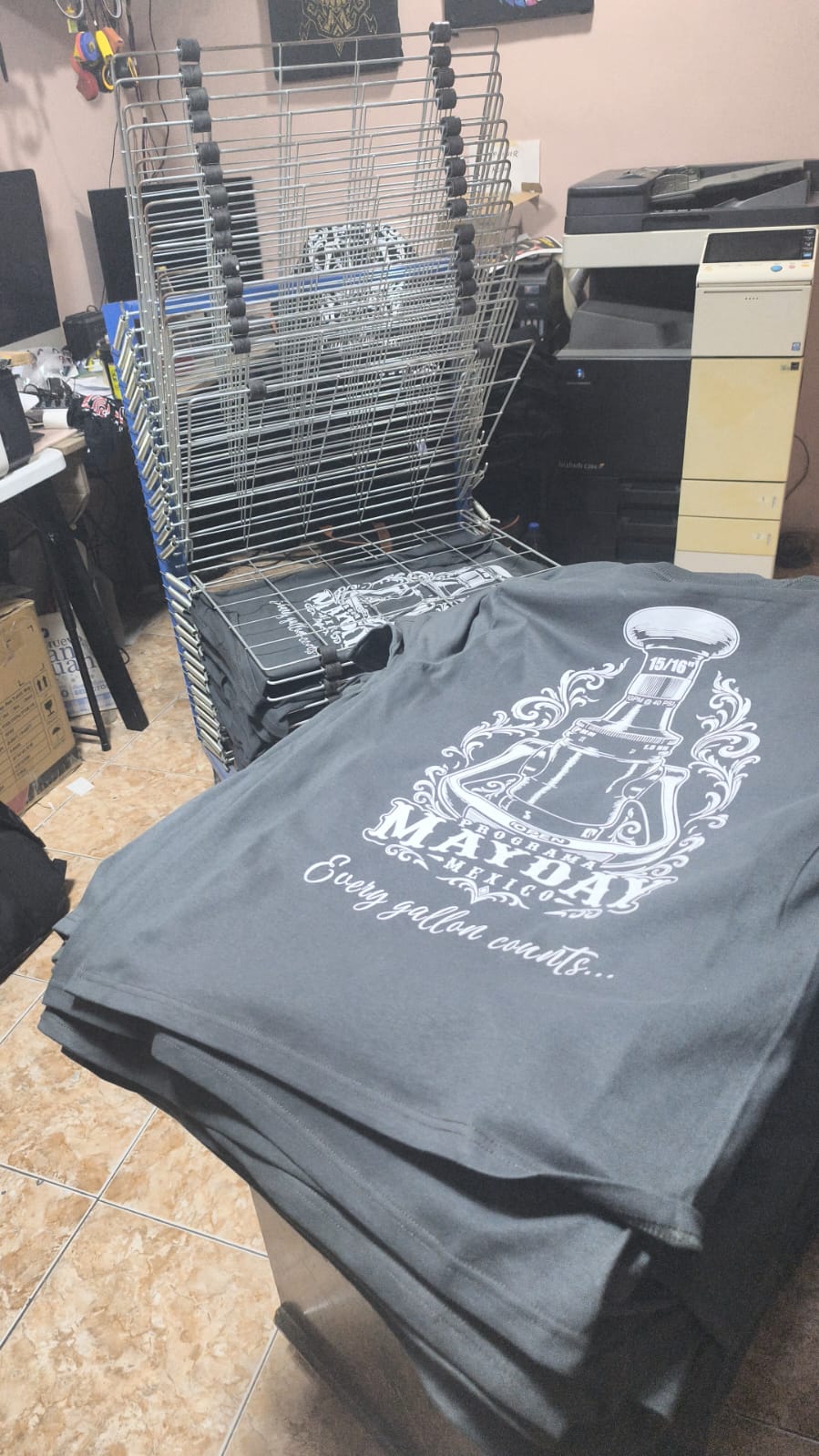 Playeras grises serigrafiadas con un diseño ornamental en tinta blanca y rack de secado en el taller